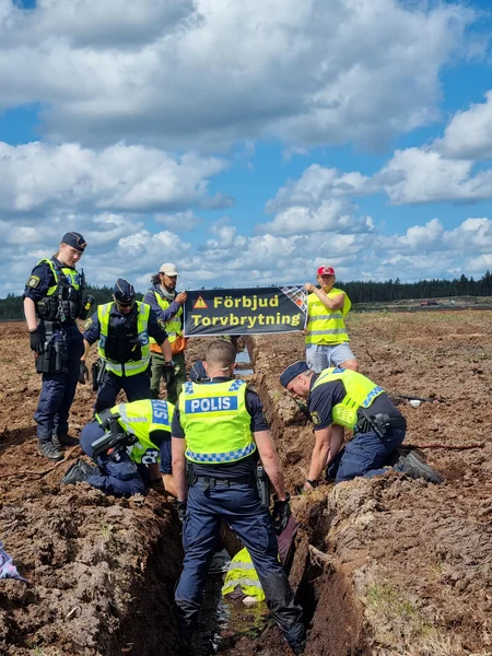 Grupp av poliser griper en aktivist från Återställ Våtmarker som ligger i en dike. I bakgrunden två aktivister med en banner "Förbjud torvbrytning"